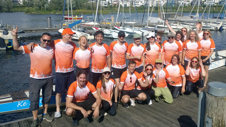 20 people in identical jerseys stand or squat on a jetty in front of a long canoe floating on the lake behind them.