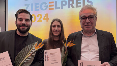 The three winners of the Faculty of Architecture stand on the Brick Award stage and hold their certificates up to the camera.