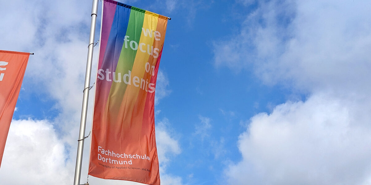 A Fachhochschule Dortmund flag with a rainbow flies against a blue sky.