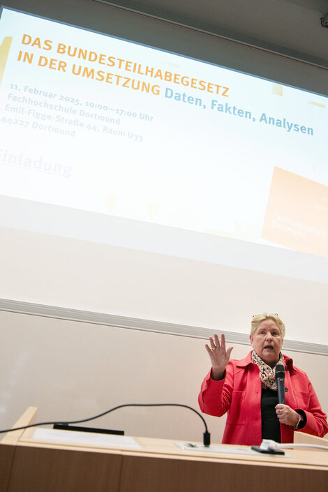 Eine Person in einem roten Oberteil steht vor einer hellen Wand und spricht, die rechte Hand gestikulierend erhoben, in ein Mikrofon. Über ihr ist an der Wand eine Projektion zu sehen mit der Überschrift: "Das Bundesteilhabegesetz in der Umsetzung. Daten, Fakten, Analysen".