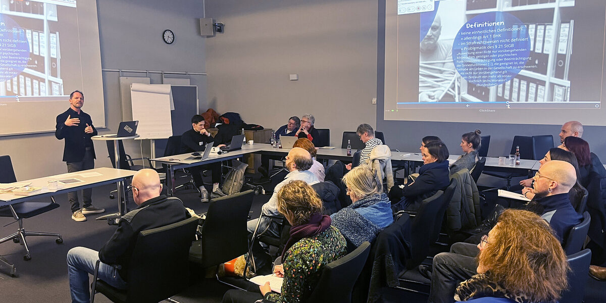 In einem großen, funktionalen Raum steht links ein Mensch an einem Stehtisch mit Laptop und gestikuliert, während ihm zugewandt im Raum verteilt 18 Personen an Tischen sitzen. Rechts und links sind Projektionen an der Wand zu sehen.