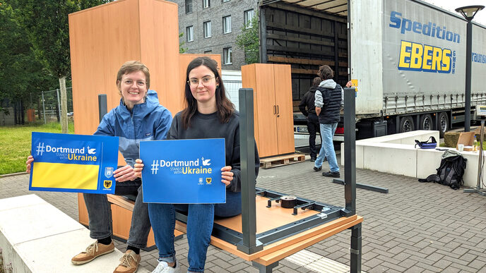 Two people are sitting on a desk holding signs reading "Dortmund stands with Ukraine". In the background, furniture is being loaded onto a truck.