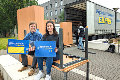 Zwei Personen sitzen auf einem Schreibtisch und halten Schilder mit der Aufschrift "Dortmund stands with Ukraine" in der Hand. Im Hintergrund werden Möbel auf einen Lkw verladen.
