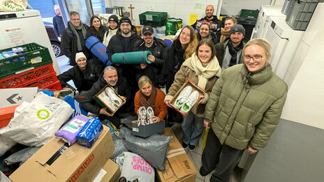 A group of people are standing in a small room full of boxes and bags. They are holding sleeping mats, sleeping bags and shoes.