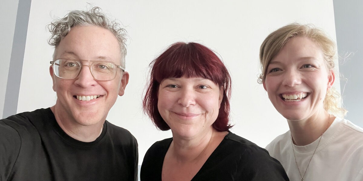 Three people in front of a gray wall smile broadly into the camera.