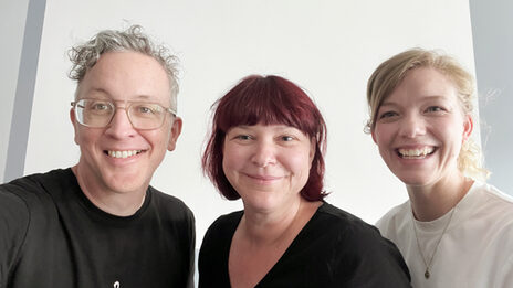 Drei Personen vor einer grauen Wand lächeln breit in die Kamera.