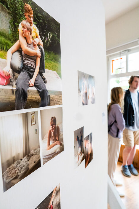 Im Vordergrund eine Bilderserie, die zwei queere Freundinnen zeigt. Im Hintergrund Gäste, die sich unterhalten.