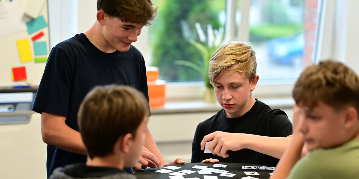 Vier Schüler an einem Stehtisch mit kleinen Karten, die Daten repräsentieren. Ein Schüler hält eine Karte hoch, während die anderen ihm zusehen.