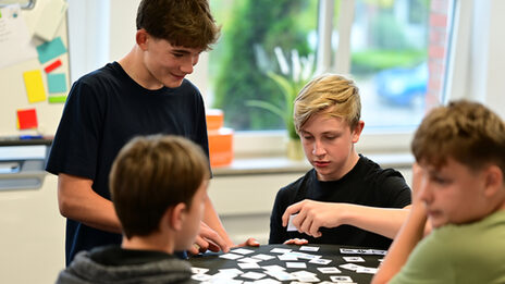 Vier Schüler an einem Stehtisch mit kleinen Karten, die Daten repräsentieren. Ein Schüler hält eine Karte hoch, während die anderen ihm zusehen.