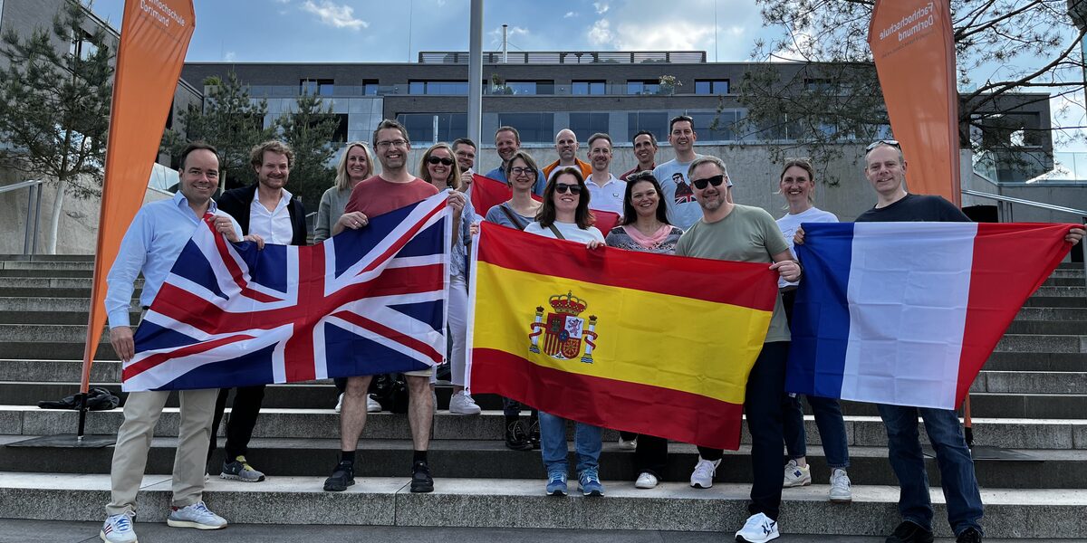 Several people are standing outside on a staircase, framed by two beach flags from Fachhochschule Dortmund. The people in the front row are holding flags of Great Britain, Spain and France in front of them.