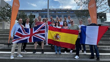Several people are standing outside on a staircase, framed by two beach flags from Fachhochschule Dortmund. The people in the front row are holding flags of Great Britain, Spain and France in front of them.
