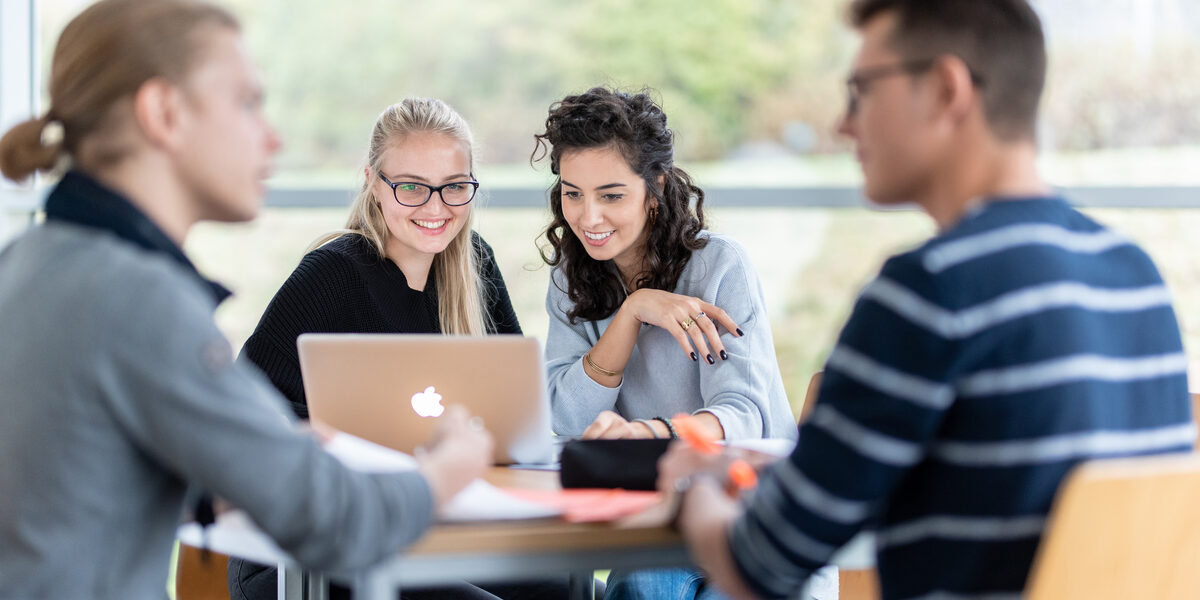 Photo of four students at a group table. The focus is on two female students sitting next to each other and looking at a laptop. Two other students are engrossed in a conversation.