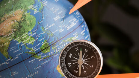 Photo of a still life: globe in the background, FH cube, orange paper airplane and compass in front of it.