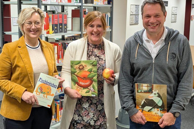 Drei Personen stehen nebeneinander, sie halten jeweils ein Buch vor sich, die Person in der Mitte zusätzlich einen Apfel. Im Hintergrund sind Regale mit weiteren Büchern zu sehen.