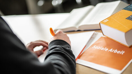 Foto einer Person im Anschnitt, die etwas mit einem orangefarbenen Stift auf einen Block schreibt. Daneben liegen Bücher und ein Heft mit Aufschrift "soziale Arbeit".