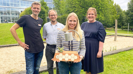 Four people are standing on the Fachhochschule Dortmund campus, one person is holding a prize trophy.