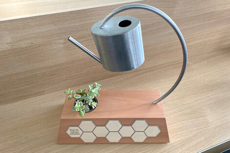 The trophy: a wooden beam engraved with the name of the prize winner and a small plant on top. A small watering can hovers above the plant. It is connected to the wooden beam via a metal arch.