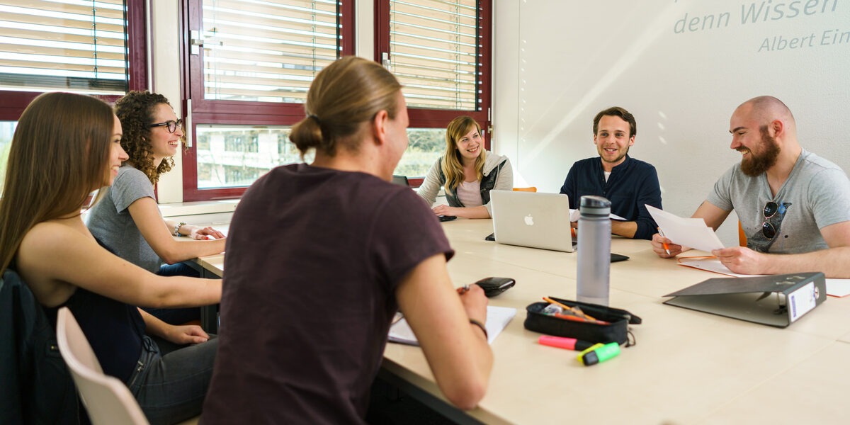 Foto von sechs Studierenden an einem Gruppentisch, die sich austasuchen. Auf dem Tisch liegen Arbeitsmaterialien.__Six students at a group table exchange ideas. Working materials are on the table.