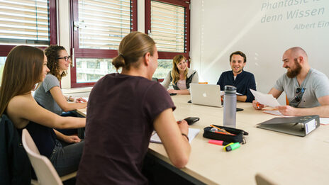 Foto von sechs Studierenden an einem Gruppentisch, die sich austasuchen. Auf dem Tisch liegen Arbeitsmaterialien.__Six students at a group table exchange ideas. Working materials are on the table.