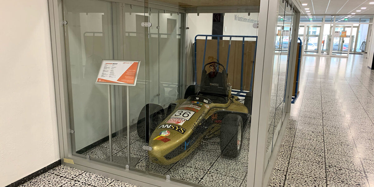 A racing car stands in a glass box next to a wall in a spacious hallway.