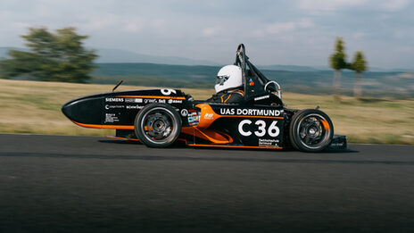 A person wearing a helmet is driving a racing car, the landscape can be seen blurred in the background.