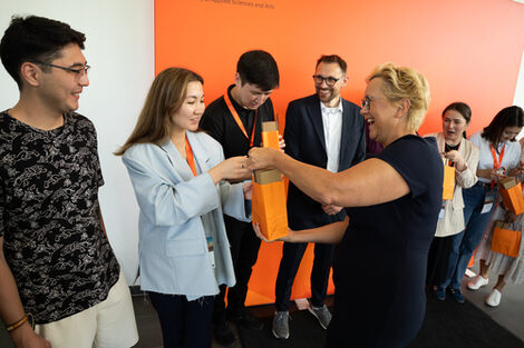 One person hands an orange, filled bag to a person on their left. Both are beaming. Five other people are standing around them.