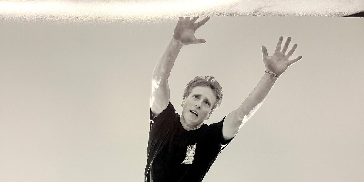 Black and white photo of a person in short, black clothing seen from below, performing a pike jump barefoot.