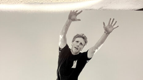 Black and white photo of a person in short, black clothing seen from below, performing a pike jump barefoot.