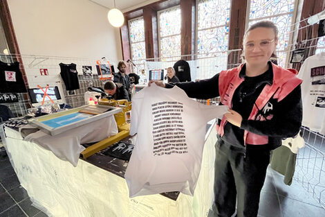 Ein Mensch in schwarzer Kleidung und roter Weste hält ein T-Shirt mit aufgedruckten Worten ins Bild. Er steht an einem Stand mit einer Siebdruckmaschine und weiteren Kleidungsstücken.