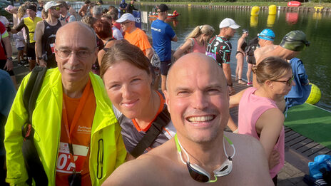 Selfie of three people under an open summer sky with many other people in the background on the bank of a watercourse.