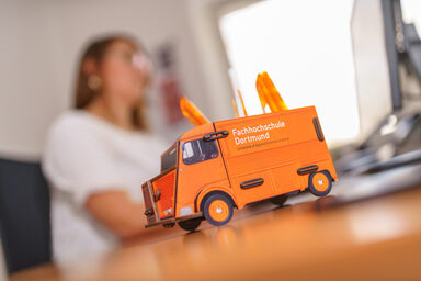 Photo of a pen box in the form of a Citroen HY panel van on a desk, in the background a woman is working on the computer. __ In the foreground on the desk is a pen box in the form of a Citroen HY panel van, in the background a woman is working on the computer.