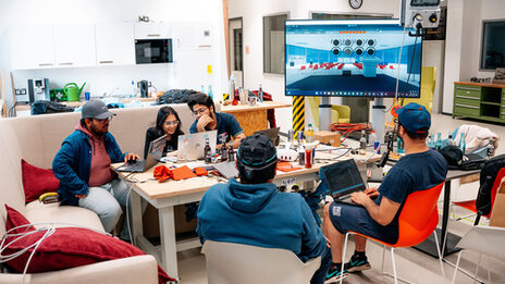 Five people sit in a room at a table with laptops and technical equipment, next to a large screen showing a virtual room with circle symbols.