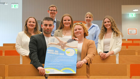 Several people are standing in the rows of seats in a lecture hall, the two at the front are holding a poster.