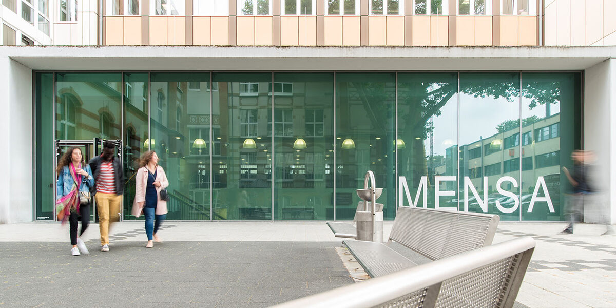 Three students come out of the new canteen on Sonnenstrasse. The glass façade of the canteen is large in the picture.