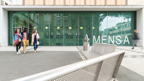 Drei Studierende kommen aus der neuen Mensa an der Sonnenstraße. Groß im Bild ist die Glasfassade der Mensa.