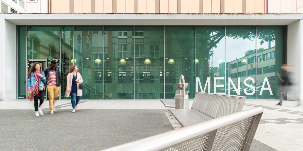 Photo of the canteen building, benches in the foreground. Four people blurred in front of the canteen __The canteen building, benches in the foreground. Four people blurred in front of the canteen.