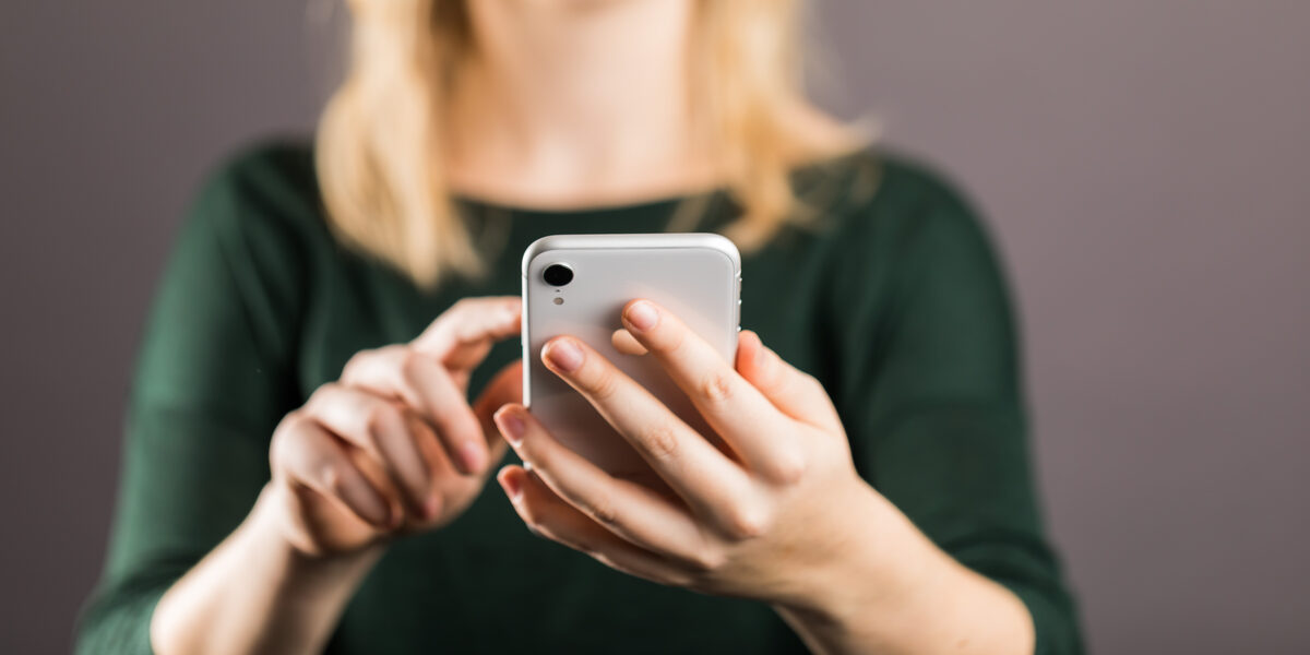 Junge Frau - Kopf oben im Anschnitt - hält vor sich ein Smartphone in der linken Hand, mit dem Zeigefinger der rechten Hand berührt sie fast das Display.