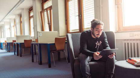 Photo of a young man sitting on an armchair and looking into his tablet. A single row of student desks is visible in the background.