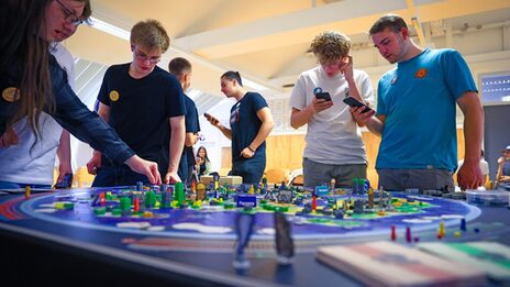 People stand around a large game board and discuss the next step in the game.