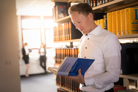 Photo of a doctoral student standing in front of a bookshelf in the library, reading a book entitled "Handbuch Promotion". Two other people in the background.