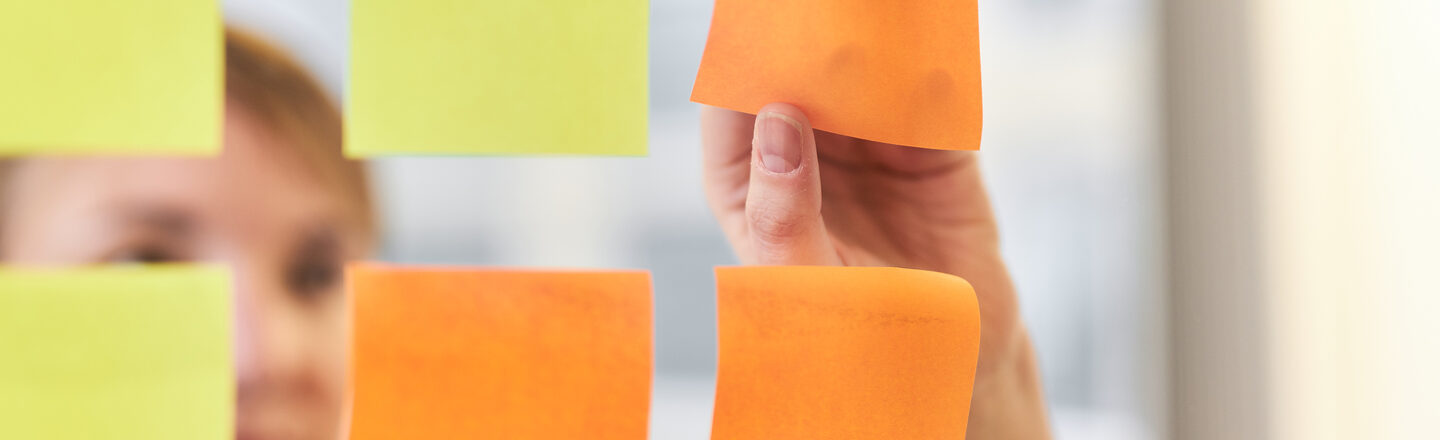 Photo of a pane with three orange and three yellow square Post-its stuck to it in two rows. Behind it is a woman pulling off an orange Post-it.