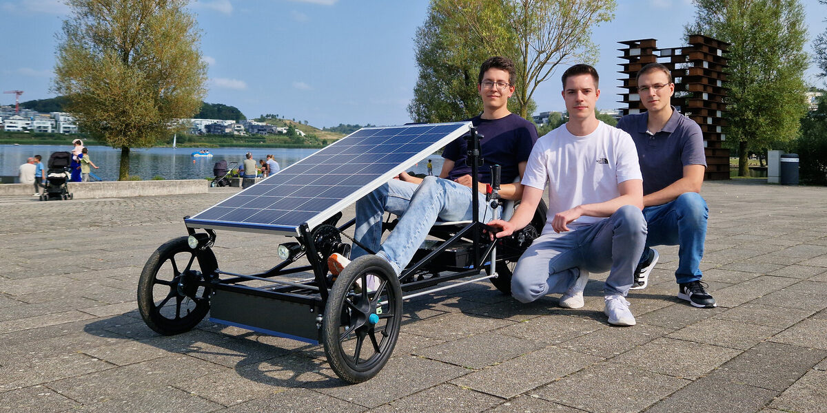 A vehicle with three wheels and a large solar panel is parked in front of a lake, one person is sitting in it and two other people are crouching next to it.