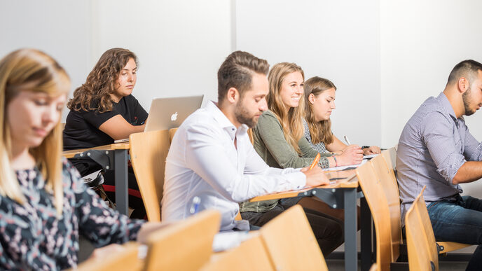 Photo in rows of seats in a lecture hall where six students are sitting at folding tables. The focus is on a smiling student, the others are looking at the tables.