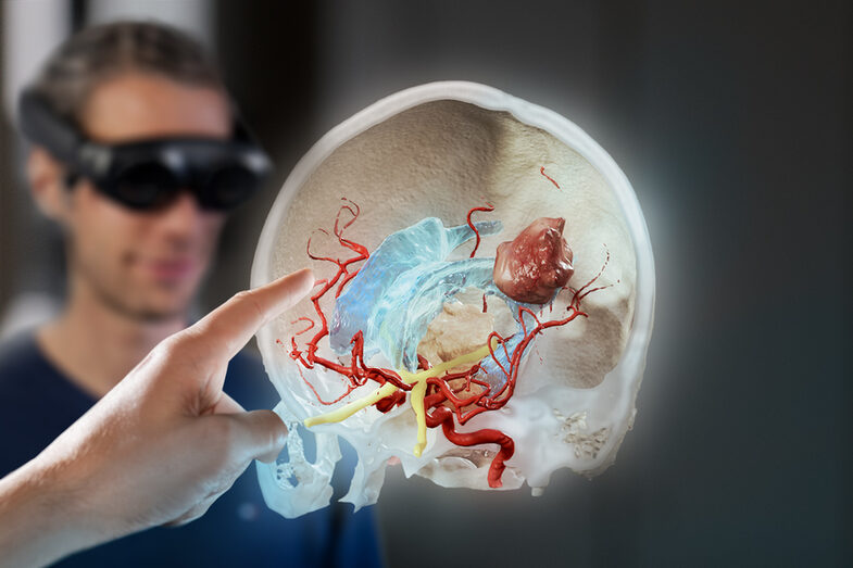 A young man in the background looks through virtual reality glasses at a cross-section of brain layers with various anatomical structures. A hand appears in the foreground on the left edge of the image, pointing at the hologram with its index finger.