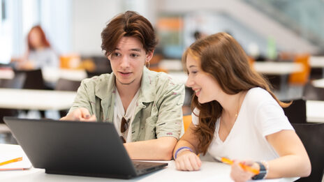 Photo of a student sitting next to each other at a table and looking at a laptop in front of them.