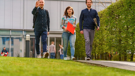 Photo of a female student and two male students walking side by side on the campus. One student waves to someone __Three students walk side by side on a path, the student on the left raises his hand in greeting.