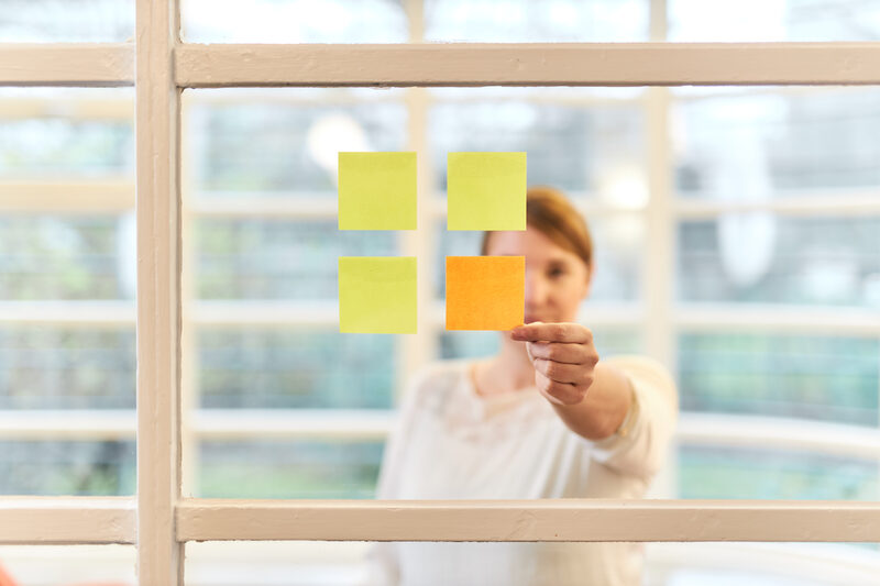 Frontal view of a woman standing behind a pane with four square Post-its stuck to it, which in turn form a square. The woman grabs the only orange Post-it.