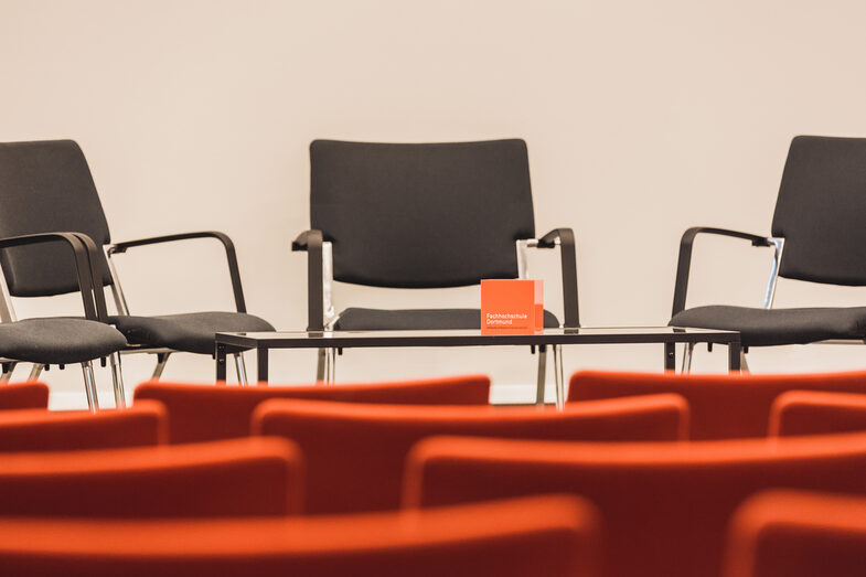 Foto eines leeren Veranstaltungsraums, Blick über leere Stuhlreihen hinweg zum Podium, wo weitere Stühle stehen sowie ein Tisch mit FH-Würfel. __ Empty event room, view over empty rows of chairs to the podium, where there are more chairs and a table with FH cube.