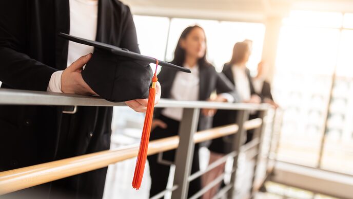 Foto von vier Promovierenden - unscharf zu erkennen - die an einem Geländer im Gebäude stehen. Im Fokus ein Doktorhut, den einer von ihnen in der Hand hält.