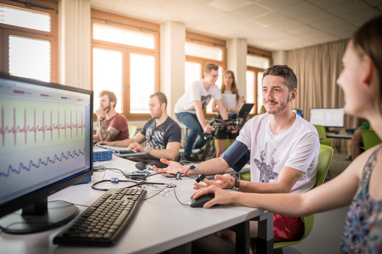 Photo of several students in a room. In the foreground, a student with a blood pressure monitor on his arm next to a student at a PC. The monitor shows the measured values. In the background is a student on an ergometer.